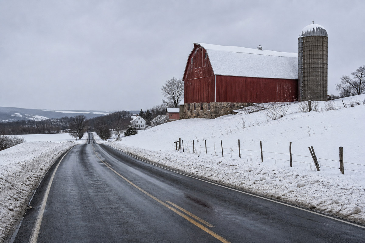A snow covered central New York dairy farm on a hillside with a red bank barn, a plowed two lane road in the foreground and a gray lake effect sky