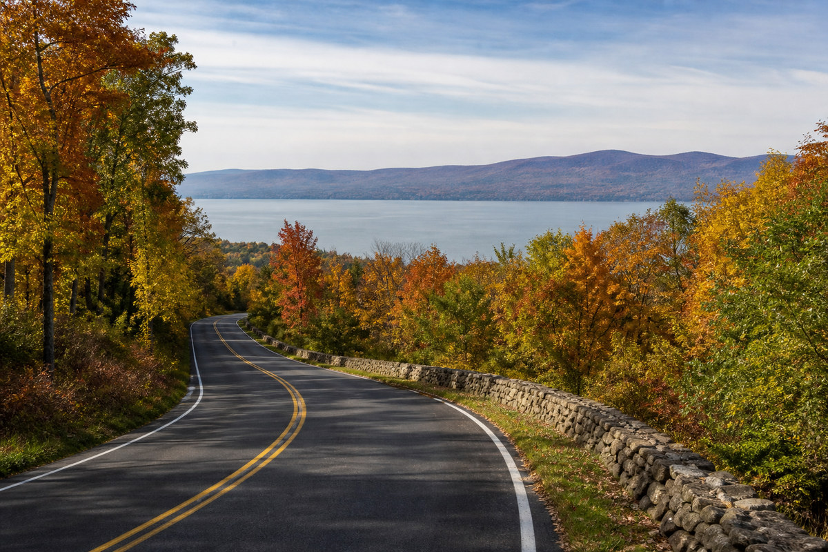 A two lane road along the west bank of the Hudson River in autumn with a stone wall on one side, hardwoods turning color and the river and the Catskill ridgeline behind
